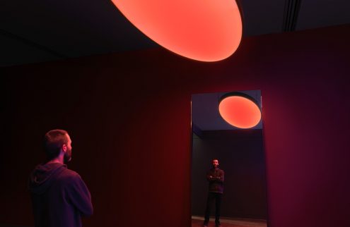 A photograph of a man standing in an installation artwork, within a red lit room and a light red orb like object appearing in the foreground, and again in a mirror reflection