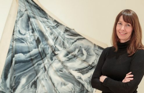 A woman stands smiling and looking directly into the camera in front of a hung textile artwork