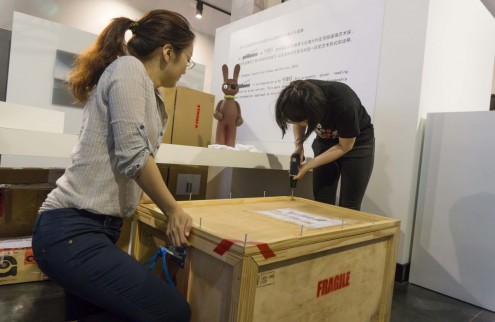 Two people are seen packing a crate. One of the people to the left of the photo holds the lid of the crate shut, while the other person to the right drills the crate together