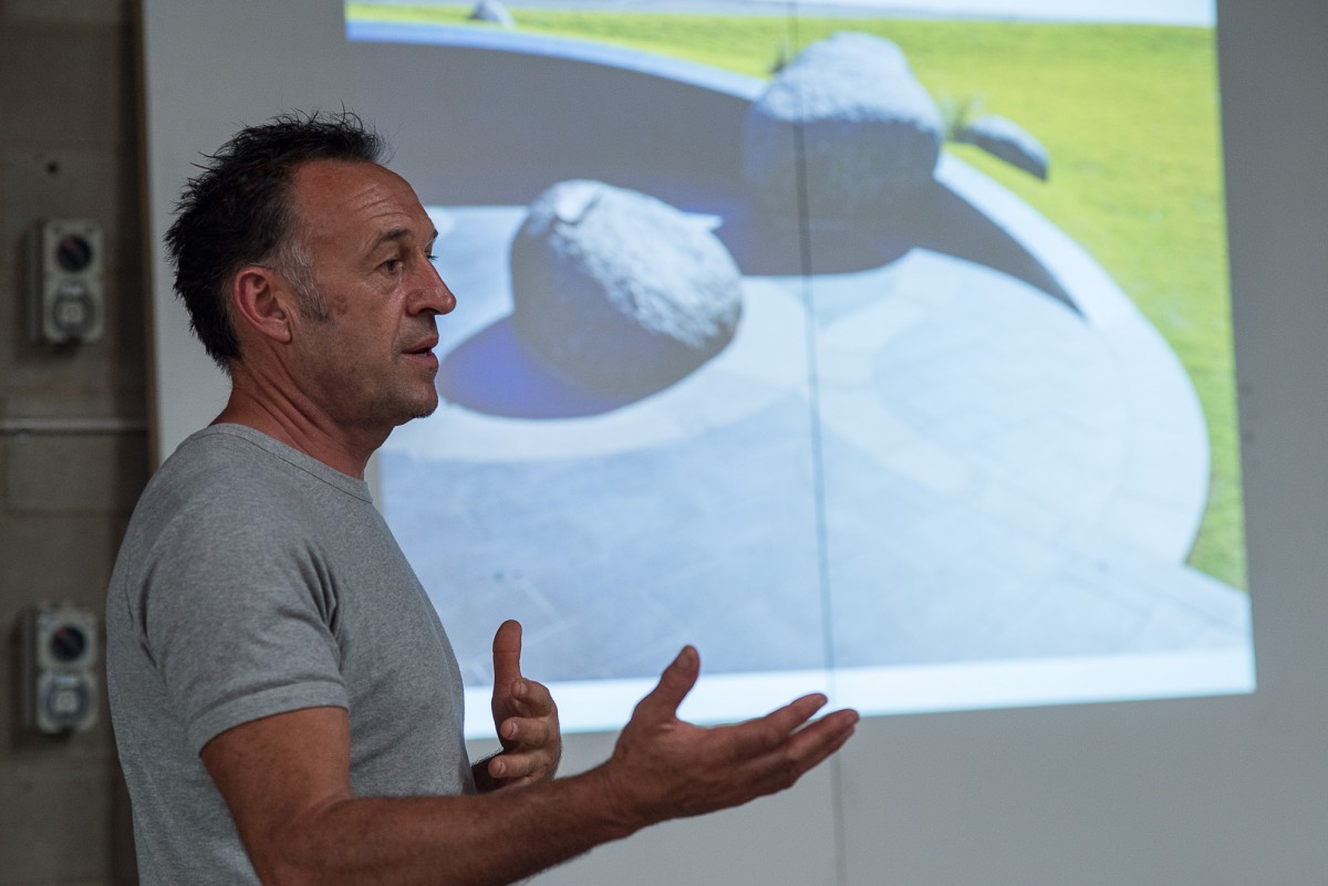 To the left of the image stands a man giving a presentation, in the background is a photograph of public art projected onto the wall