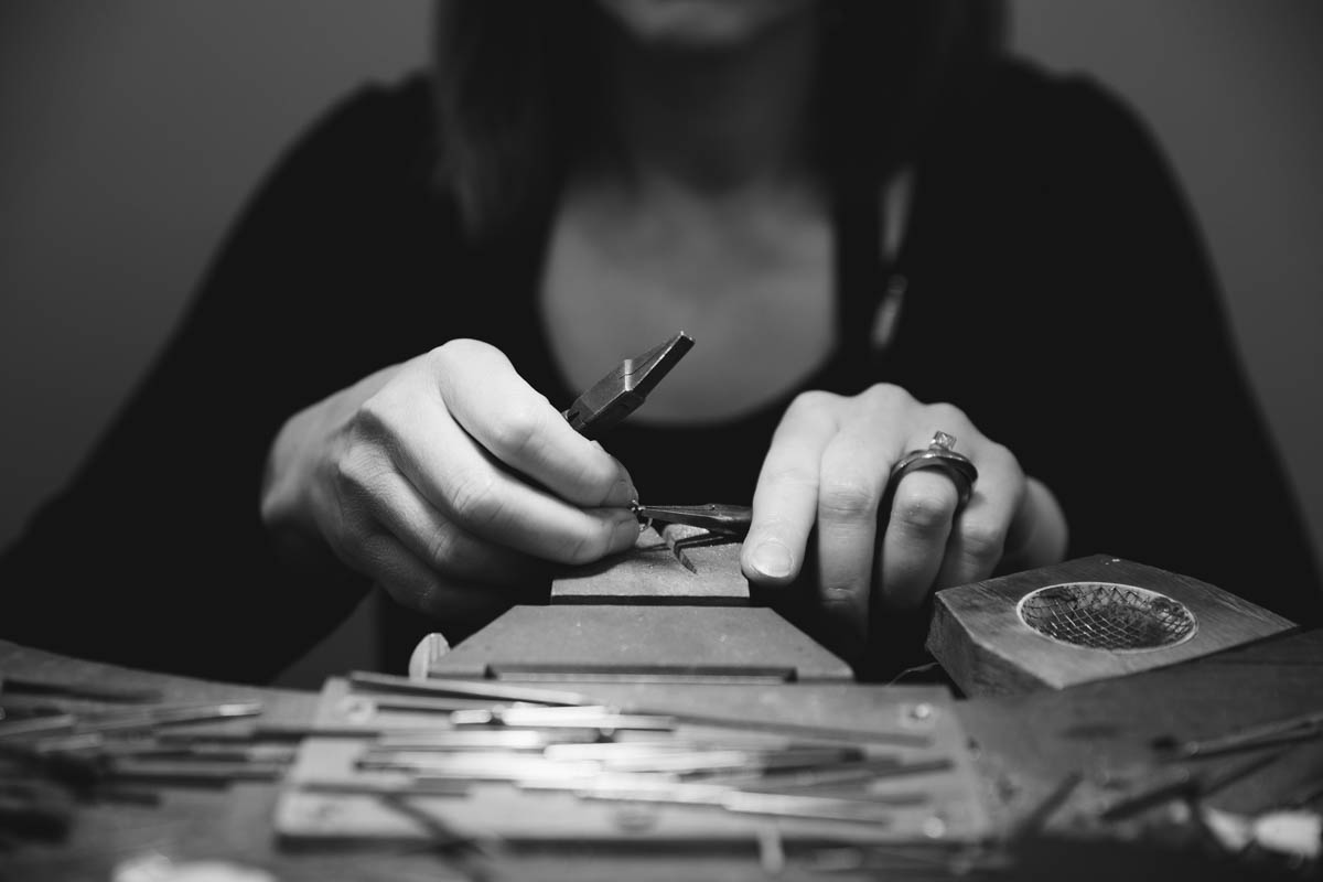 A close up and detailed black and white photograph of a person creating metal work by hand. The person holds a pair of pliers in each hand and shapes a thin piece of metal