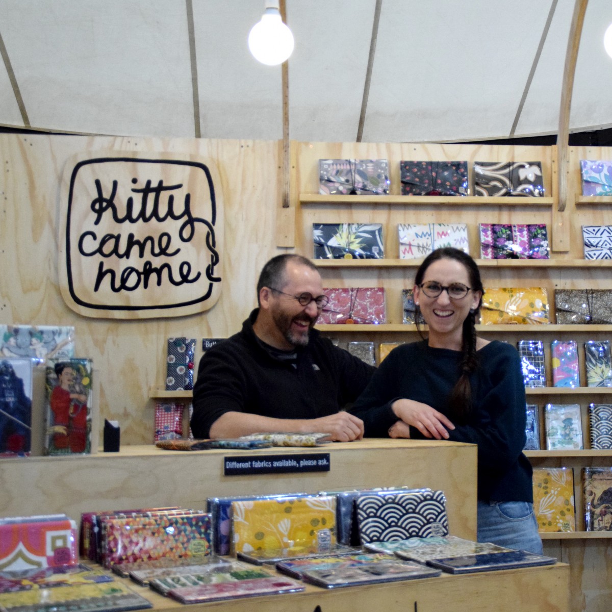 A photograph of a man and woman smiling, looking directly into the camera. In the background and foreground are collection of differently styled wallets and purses, In the left corner is a logo which reads' Kitty come home'