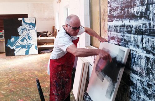 A man in a studio space stands centre of the image, and is seen painting a nearly half blank canvas