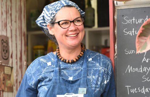 A woman stands smiling in the centre of the photograph, next to her is a chalkboard with dates of the week written on it