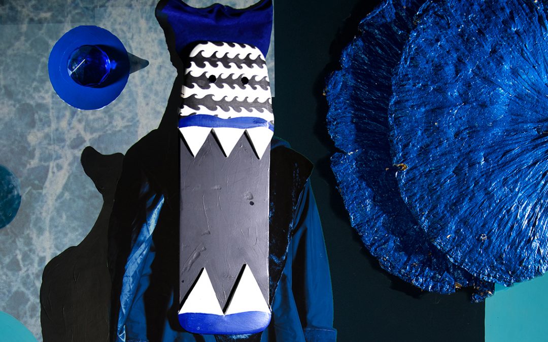 Figure wearing blue costume and elongated facial mask standing amongst blue objects.