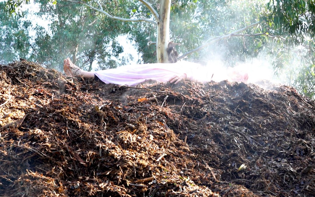 Woman in pink dress lies on top of pile soil and compost. Her face is obscured by steam or smoke.